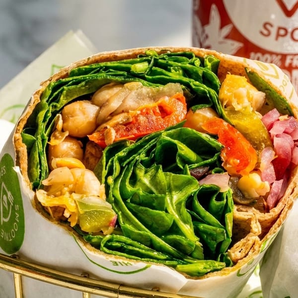 A colorful wrap filled with chickpeas, spinach, and vegetables, in a gold basket with a Smoothie Spot cup.