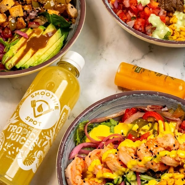 Three colorful salad bowls with various toppings and two bottles of Smoothie Spot Tropical Juices.
