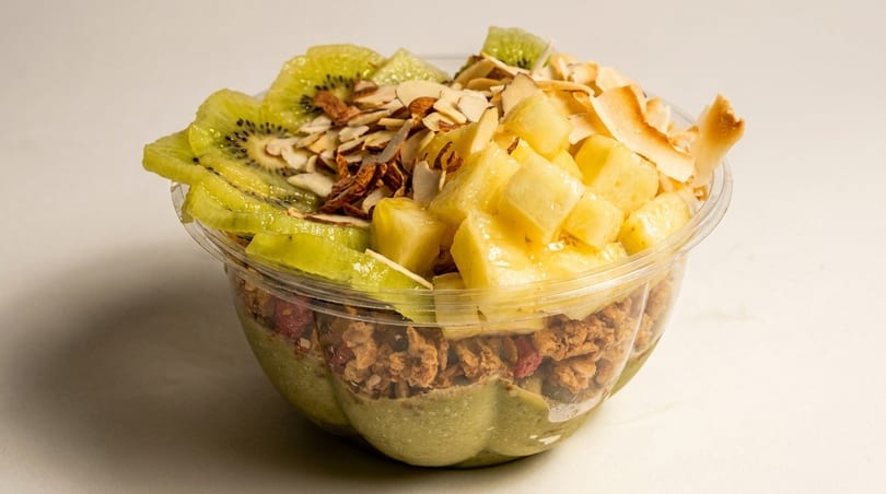 A vibrant smoothie bowl with kiwi, pineapple, almonds, and granola in a clear cup.