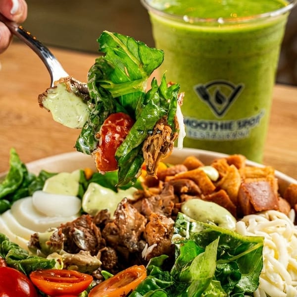 A hand holding a forkful of salad with chicken, tomatoes, and dressing, next to a green smoothie.