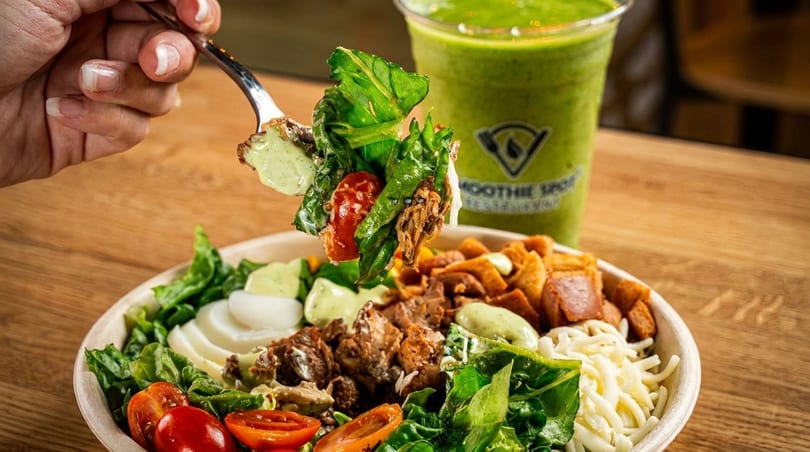 A hand holding a forkful of salad with chicken, tomatoes, and dressing, next to a green smoothie.