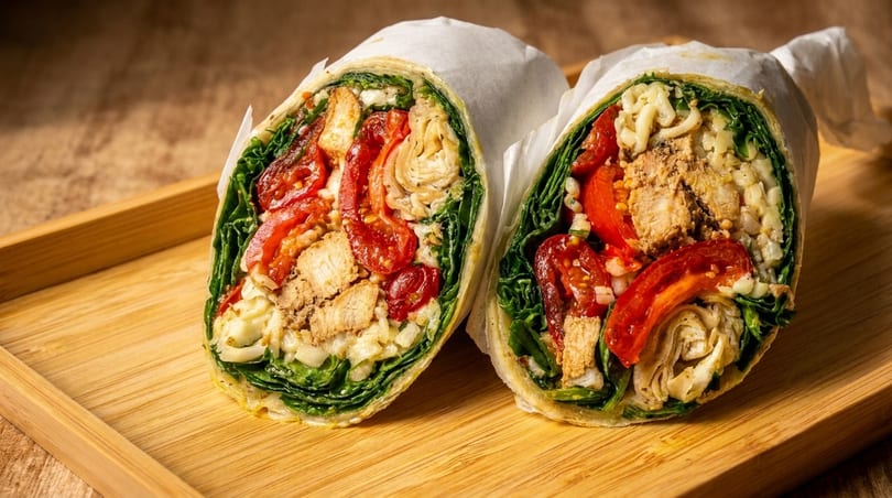 Two halves of a chicken and vegetable wrap, filled with greens, tomatoes, and rice, on a wooden tray.